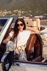 Young woman in a convertible car in front of Lake Como with a Trolley and a Legacy Bag from the Bellagio collection in Cream color. | Bric's Milano